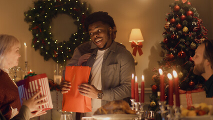 Happy multi cultural family celebrating Christmas or New Year 2023, exchanging gifts. Served holiday table with different dishes and candles. Warm atmosphere of family Christmas dinner at home.