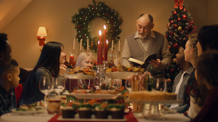 Drandfather reads Bible on family Christmas dinner. Large diverse family praying before celebrating Christmas or New Year 2023. Served holiday table with delicious meal and candles. Winter holidays.