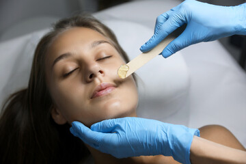 Young woman undergoing hair removal procedure on face with sugaring paste in salon