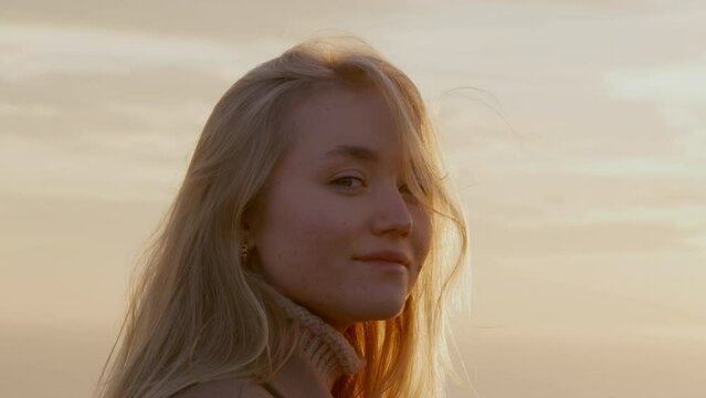 Portrait of a young woman against picturesque bay with soft, sunset lighting. Close-up shot of a blonde-haired girl with cheerful facial expression. High quality 4k footage