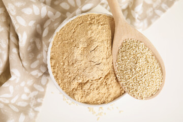 Sesame flour in bowl and wooden spoon of seeds on white table, top view