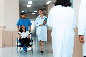 Doctor and male nurse transport a female patient in a wheelchair along sterile hospital corridor....