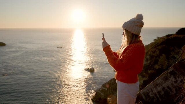 Spectacular view of the ocean, washing rocky cliffs with calm waters. Woman taking pictures of postcard, panoramic scenery with sunset skyline in the background. High quality 4k footage