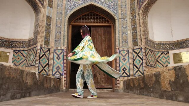 4K. Overall plan. Tourism in Uzbekistan. An oriental girl in a traditional Uzbek robe dances with a robe against the backdrop of an old madrasah in Bukhara. Slow motion.