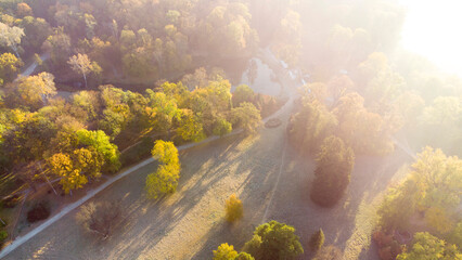 Aerial flying over trees with yellow leaves, meadow, dirt road with morning mist an autumn sunny morning in park. Bright sunlight, shining sunbeams rays, sun overexposure. Beautiful natural background