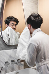 Obraz premium Asian man wearing bathrobe is washing face with towel in front of the mirror.