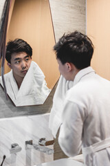 Asian man wearing bathrobe is washing face with towel in front of the mirror.