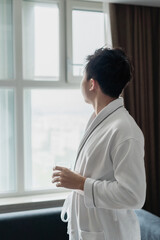 Asian man holding a glass of water is standing near the window.