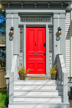 Grey wooden steps led up to a bright red metal door. There's a mail slot in the door, a black handle, two flower pots on the step, with black light fixtures, and a grey exterior wall of a house. 