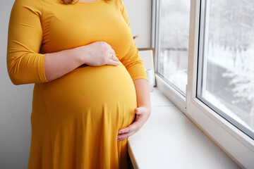 A pregnant woman in a yellow dress strokes her belly and poses near the window. It's winter outside and it's snowing. Close-up of the belly