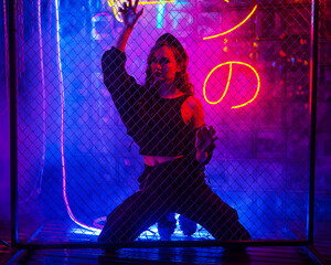 Caucasian woman in neon studio behind chain-link mesh. 