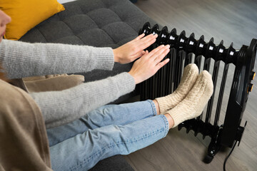 Women sitting and warming hands over and electric heater. High energy prices and cold home.