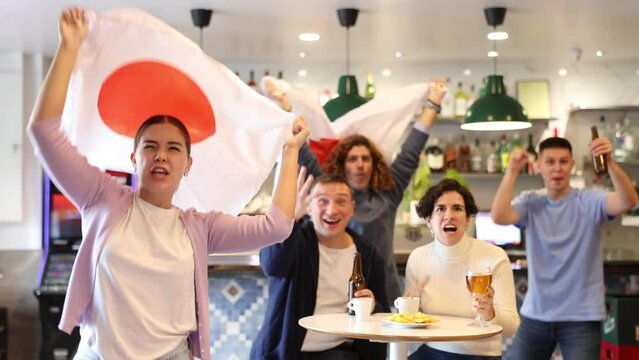 Group of cheerful emotional people of different ages and nationalities cheering for favorite sports team in pub, waving national flag of Japan while watching match on TV. High quality 4k footage