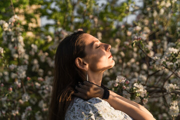 Attractive woman with closed eyes enjoying fresh air and sun in blooming apple tree garden. She holding hair with arms.