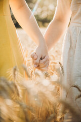 Two unrecognizable teenage caucasian girls wearing dresses holding hands in golden wheat field in sunny summer day