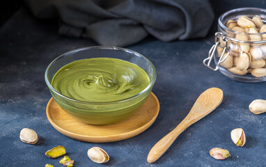 Pistachio spread cream in glass bowl raw pistachios in jar, napkin, wooden tea spoon on dark stone...