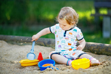 Cute toddler girl playing in sand on outdoor playground. Beautiful baby in summer clothes having fun on sunny warm summer day. Outdoors activity for toddlers.