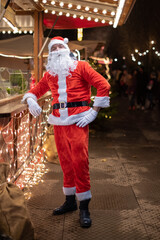 Portrait von einem verkleideten Weihnachtsmann, der auf einem Weihnachtsmarkt von Lichtern umgeben...