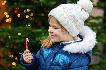 Little cute preschool girl with candy cane from a sweets stand on Christmas market. Happy child on traditional family market in Germany. Preschooler in colorful winter clothes during snowfall