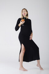 pregnant young woman with a big belly in a black dress poses with a glass of carrot juice