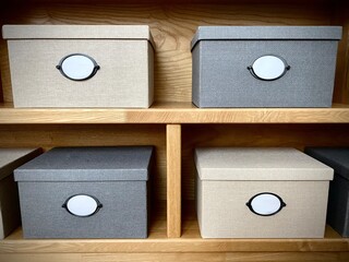 close-up of boxes with blank white labels stacked in a row on top of each other. Lots of storage boxes made of fabric in dark and light colors on a wooden shelf. Home box storage system close up.