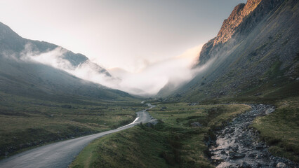 Clouds roll over Honister Pass after sunrise. Beautiful landscape scenery in Lake District, UK. Cloud inversion in a mountain valley with a road and stream.