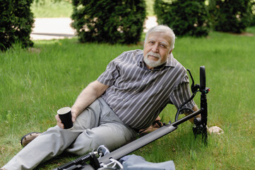 A positive, elderly man went for a walk to the park, stopped and lay down to rest on the grass next to an electric scooter. Summer, sunny day. Healthy lifestyle of the elderly.