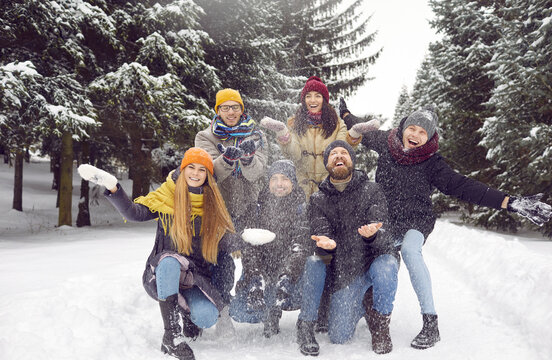 Happy young millennial friends having fun in winter park. Cheerful adult people in warm hats, coats and jackets throwing snow in air while posing for funny group photo on snowy road among fir trees