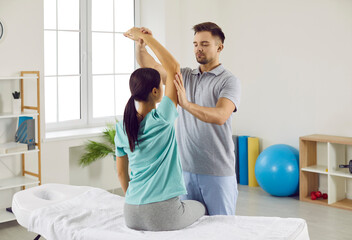 Male chiropractor, physiotherapist or osteopath at modern therapy and rehabilitation center checking womans arm. Physiotherapy specialist working with female patient whos sitting on examination bed - obrazy, fototapety, plakaty