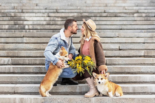 A Young Couple In Love On A Date In The Park. A Blonde In A Hat And A Man Look At Each Other. Walking With Corgi Dogs. Spring