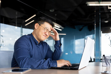 Asian businessman boss sick has severe headache, man in glasses sitting at desk using laptop at work inside office, migraine office worker investor overtired.