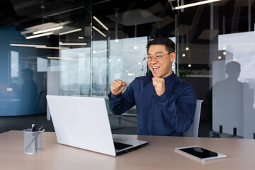 Successful businessman celebrating victory, man dancing sitting at table, asian man in shirt and glasses at work inside office with laptop, satisfied with achievement investor.