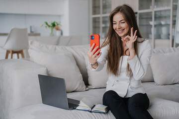 Cheerful Italian young woman makes video call by phone sitting on couch with laptop at home gestures ok sign toothy smiles happy by new home. Satisfied successful businesswoman at distant meeting.