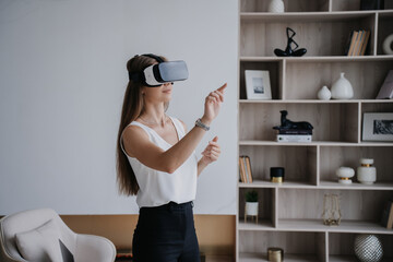 Young fit caucasian woman in white blouse and black pants using vr googles at home, playing game. Pretty Italian student girl educates by virtual reality headset. Remote education and Entertainment. - obrazy, fototapety, plakaty