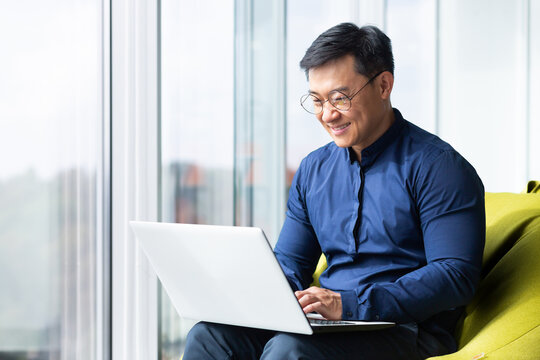 Successful and smiling asian man working with laptop inside office with notebook, senior businessman in glasses and shirt.