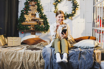 Cute young woman talking on phone to prepare for Christmas with laptop and technology while sitting on bed near Christmas tree at home on holiday.