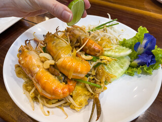 Hand of woman squeezing lemon on Pad Thai with king prawns and cucumber slices. Traditional food plate from Thailand