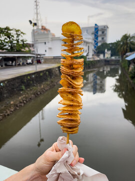 Hand of woman holding crispy potato chips on spiral skewer by the river. Traditional Thai snack concept
