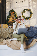 Obraz premium Beautiful Merry Christmas woman drink a sweet cup of hot cocoa, wear comfortable home clothes, look at the laptop, work remotely for the new year, plaid, New Year's winter mood.