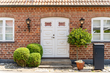 Image of White wooden vintage entrance door with metal side lamps, brick walls and green bushed in front of it