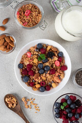 Crispy muesli with fresh berries, almonds in bowl and milk in jug, chia seeds for making pudding on light gray background. Healthy breakfast concept