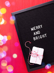 Christmas postcard. Letterboard with phrase merry and bright and box with present oh bright red paper textured background. Top iew. Bokeh light.