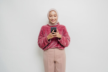 Fototapeta premium A portrait of a happy Asian woman wearing a pink sweater and hijab, holding her phone, isolated by white background