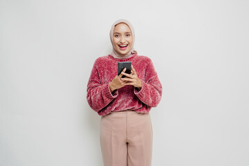 Fototapeta premium A portrait of a happy Asian woman wearing a pink sweater and hijab, holding her phone, isolated by white background