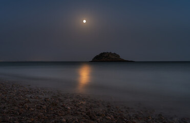 Moonlit beach with pebbles on foreground and a limestone sea stack on background in the sea, on a clear night. Colour photo, nature reserve, Serra da Arrábida, Setúbal, Portugal