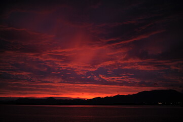 Fototapeta na wymiar Red, pink, orange and violet sunrise at the seaside during dawn with clouds in the sky and mountains on the shore of a port