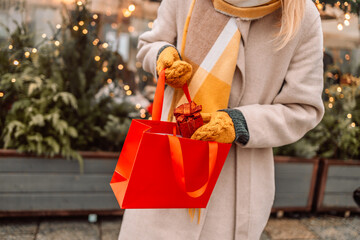 Happy young winter woman with christmas gift. Gift Box Outdoors. Surprise. Cheerful attractive girl shopper with paper bags and presents. Christmas shopping in mall. Winter sales