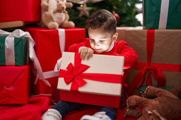 Obraz na płótnie Canvas Adorable hispanic toddler unpacking christmas gift sitting on floor at home