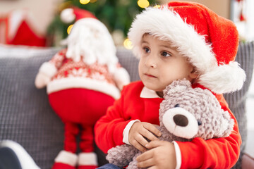 Fototapeta na wymiar Adorable hispanic toddler hugging teddy bear sitting on sofa by christmas tree at home