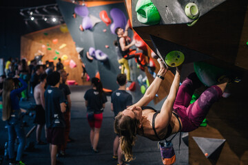 A young, slender, athletic climber in a sports field climbs a boulder at a festival in a climbing center. A vertical frame.